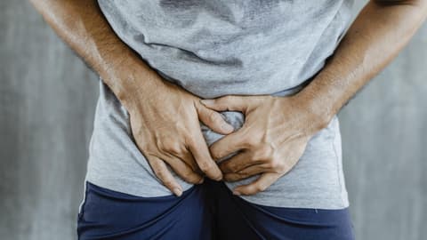 Eine Vorhautentzündung kann schmerzhaft sein Nahaufnahme eines Mannes in kurzer blauer Hose mit grauem Shirt, der sich vor Schmerzen in den Schritt greift