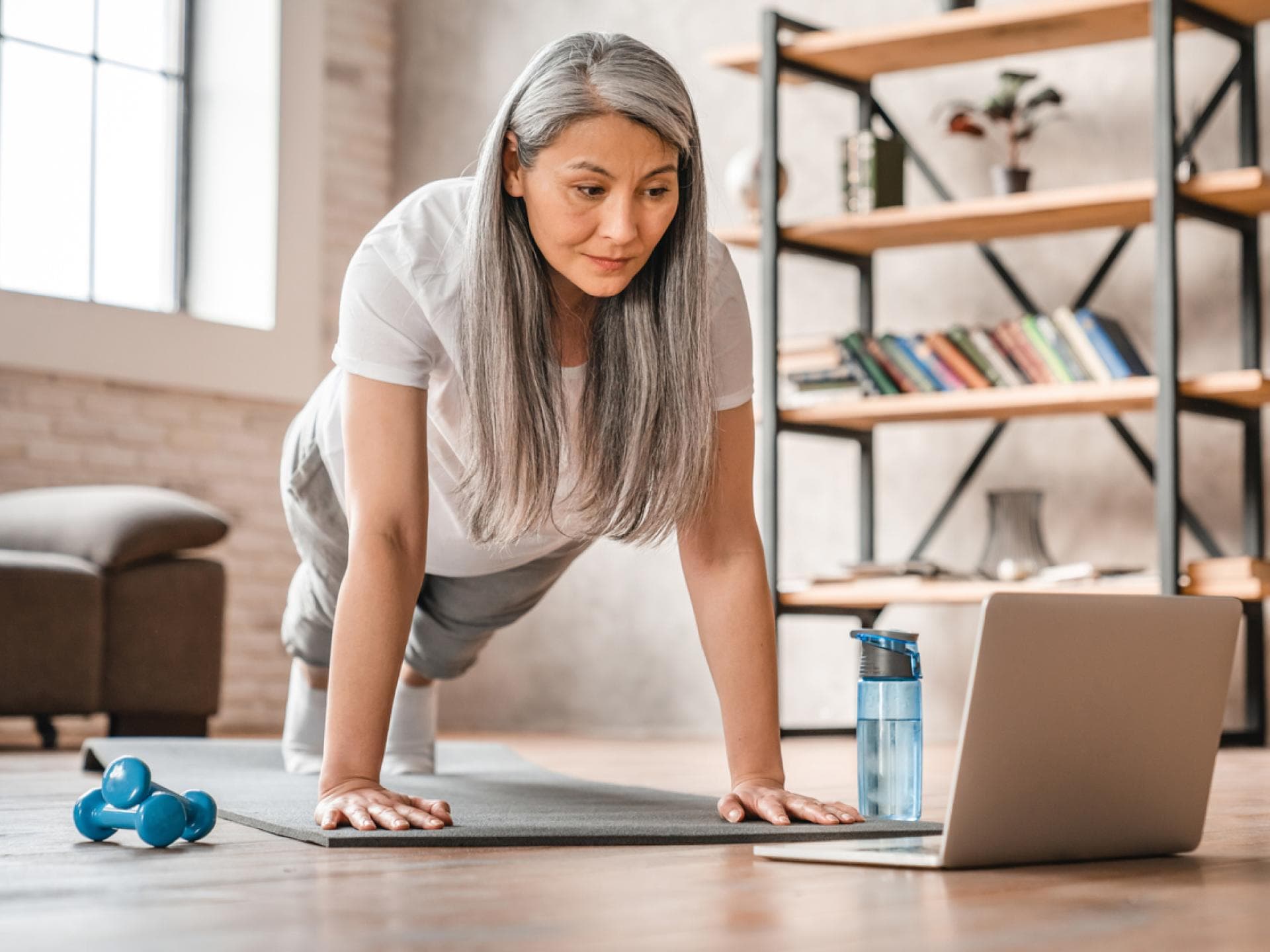 Regelmäßige körperliche Aktivität fördert den Knochenaufbau und wirkt Osteoporose entgegen. Frau im mittleren Alter macht Liegestütz zu Hause vor Laptop