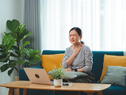 Mit der Ernährung können Sie Einfluss auf Heuschnupfen nehmen Frau sitzt im Wohnzimmer am Laptop und fasst sich an den schmerzenden Hals