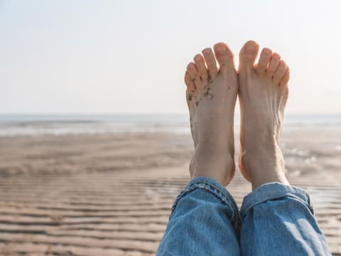 Unsere Füße werden oft unterschätzt! Darum sollten wir uns mehr um sie kümmern! Eine Frau hält ihre nackten Füße hoch. Sie liegt am Strand. Ihre Fußnägel sind naturbelassen.